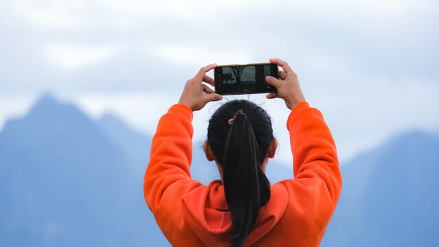 Rear view of a young woman standing on the top of a calm mountain and taking pictures of the beautiful mountain scenery at sunrise on the phone.