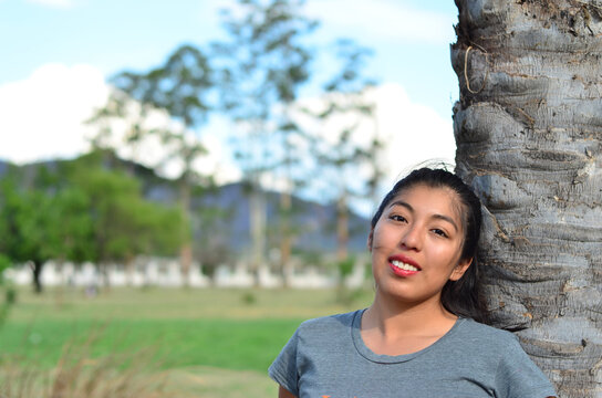 Joven latina sentada al lado de un &aacute;rbol de palmera en el parque sonriendo. Mujer morena alegre descansando