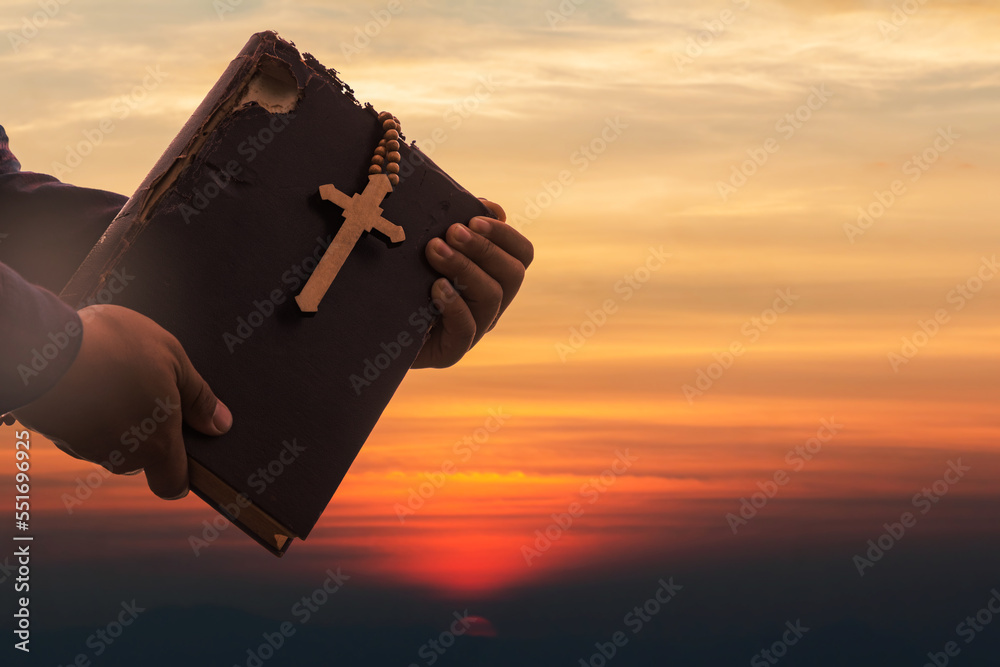 Wall mural Hands holding Old Holy Bible book and Cross on sunset sky. Religion and Hope Concept.