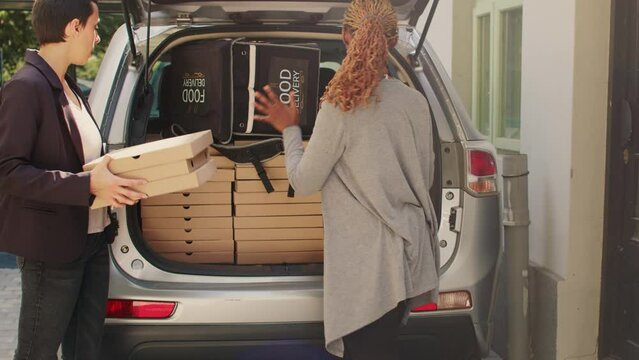 Pizzeria delivery person giving takeaway meal boxes from trunk, delivering piles of pizza packages to woman at front door. Restaurant employee taking big food order to deliver to customer.