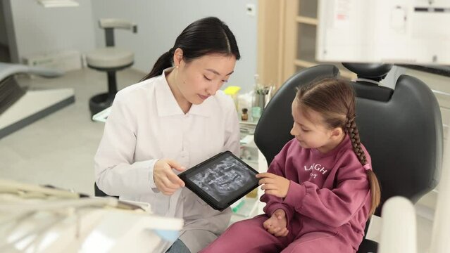 Smiling happy caucasian child girl visiting dentist, sitting in dental chair at modern light hospital clinic. Young asian woman dentist holding tablet pc with x ray scan.