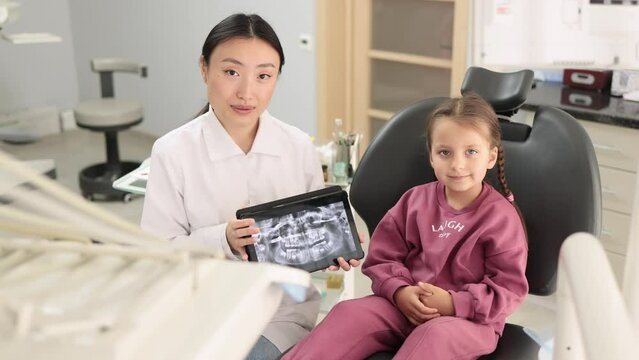 Medicine, pediatric dentistry and oral care concept. Happy female asian dentist and caucasian kid girl, looking at camera while holding tablet pc computer with x ray image.