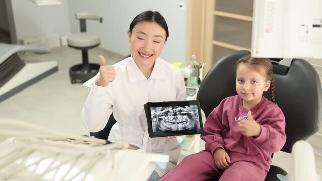 Medicine, pediatric dentistry and oral care concept. Happy female asian dentist and caucasian kid girl, looking at camera while holding tablet pc computer with x ray image showing thumb up.