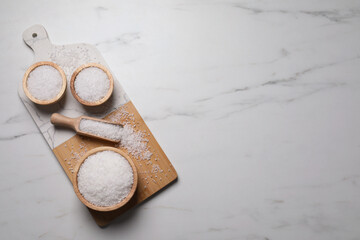 Wooden board with natural sea salt in scoop and bowls on white marble table, flat lay. Space for text