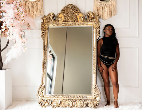 Black woman standing next to a full length mirror on a fur rug in a black body suit