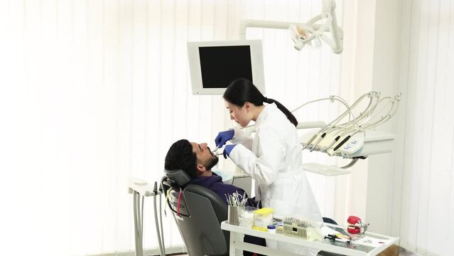 Beautiful smile with white teeth. Asian dentist treating teeth of a young bearded man using tooth drill through a magnifying glass in the dental office.