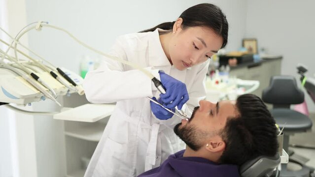 Happy patient and dentist concept. Young beautiful asian female stomatologist treating teeth of a handsome bearded young guy, using tooth drill. Oral health and hygiene.