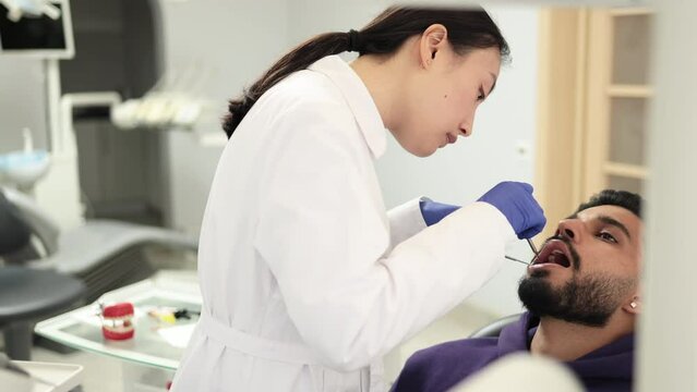 Side view of young pretty asian doctor in white uniform and gloves, making examination or curing caries and toothache for her male patient, attractive bearded man sitting in dental chair.