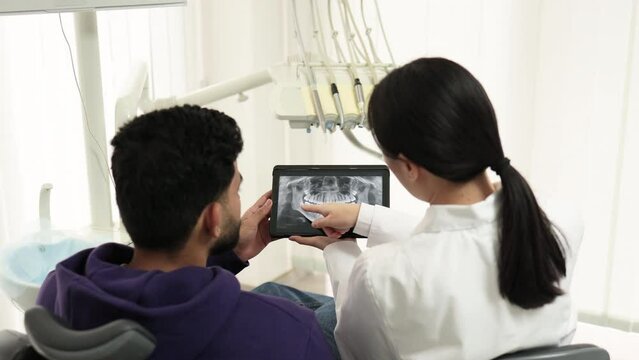 Back view of young attractive man visiting dentist, sitting in dental chair at modern light hospital clinic. Young asian woman dentist holding tablet witn x ray image. Focus on picture.