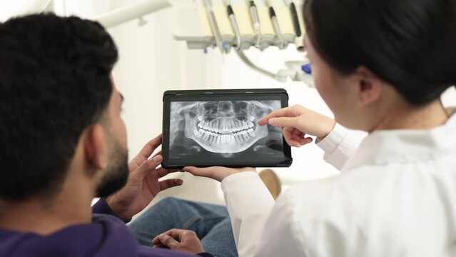 Medicine, dentistry and oral care concept. Female young asian dentist holding tablet with x ray picture with attractive patient, bearded man at modern light dental clinic.