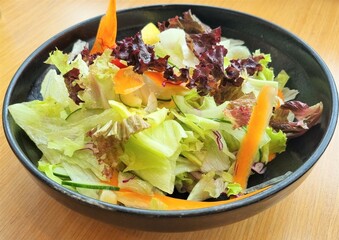 Green salad with carrot and fresh vegetables in a black bowl.