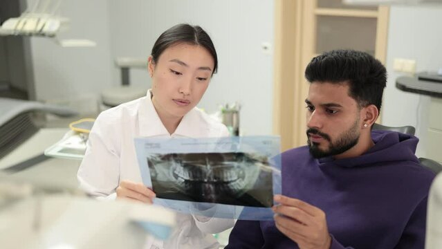 Attractive young bearded man, sitting on dentist chair and looking at x-ray picture of scan image of teeth together with his cheerful female asian dentist at clinic.
