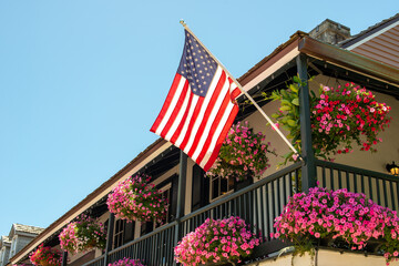 American flags hang off a second floor balcony of a historic building. The railings have baskets of pink and purple flowers hanging. The sky is blue and there are multiple windows in the old building.