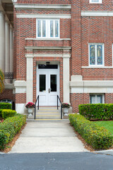 The entrance to a red brick building with tall clear glass windows, and double glass metal doors. There are concrete steps and green shrubs leading up to the facade of the commercial business property