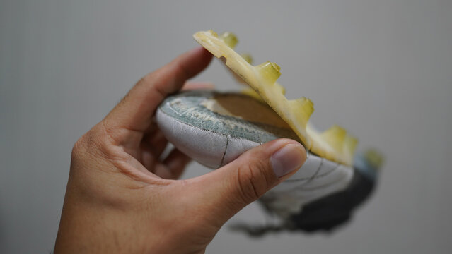 A people hand is examining a broken football shoe's sole which is parted from the upper body due to very old. Sport equipment object, close-up and selective focus. 