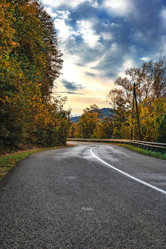 Highway on an autumn day among the mountains under a blue cloudy sky. Yellow-orange foliage on trees near the highway. An asphalt road with fallen leaves in an autumn forest. Vertical image. 