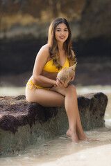 Young Asian woman sitting on the rock, holding coconuts. Summer beach concept. Tanned skin. Beautiful woman wearing yellow swimsuit. Travel to tropical island. Suluban beach, Bali
