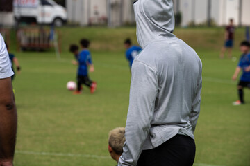 Obraz premium Dad standing and watching his son playing football in a school tournament on a sideline with a sunny day. Sport, outdoor active, lifestyle, happy family and soccer mom and soccer dad concept.
