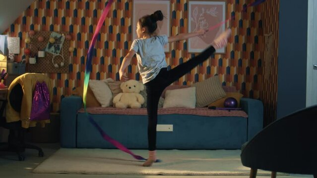 Young asian girl dances with gymnastic ribbon, showing vertical leg-split and spins around herself. Gymnastics training. Sports hobby. Upgrading physical health on quarantine. Slow motion.