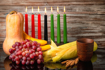 Kwanzaa holiday concept with traditional lit candles, pumpkin, corns, grapes, banana, bowl and of wheat with reflection on black background