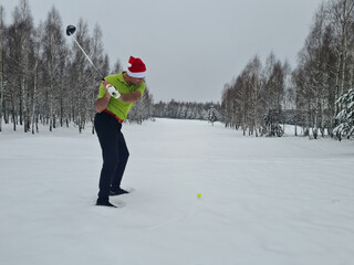 Male golfer in santa claus hat in winter on golf course with golf clubs hitting ball