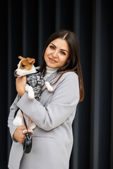 Woman in casual jacket hugging her favorite fluffy trained purebred Jack Russell Terrier dog dressed in suit for dogs near wall in outdoor in autumn day.