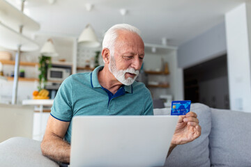 Fototapeta premium Mature man using laptop, holding plastic credit or debit card, senior grey haired customer making secure internet payment, shopping or browsing online banking service, entering information