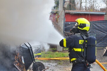 A firefighter with a breathing apparatus uses water to extinguish a fire in a wooden structure with thick smoke and flames