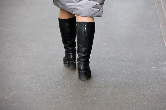 Female Legs In Black Boots. Woman In Warm Coat Walking On Winter Street