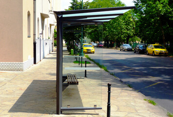 bus shelter of glass and aluminum structure in side view. urban street with car traffic and lush...