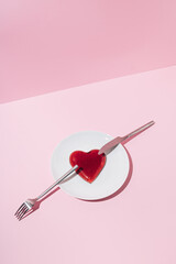 Red jelly heart, silver knife and fork on white plate and pink color background. Creative love or Valentines minimal concept.
