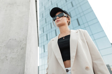 Beautiful fashionable urban woman with stylish sunglasses and a black cap in fashion casual clothes with a blazer and top walks in the city