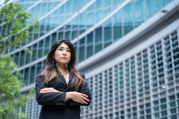 Fototapeta premium Confident Asian business woman with crossed arms for her achievements, modern skyscrapers background, female businesswoman of Japanese origins, copy space