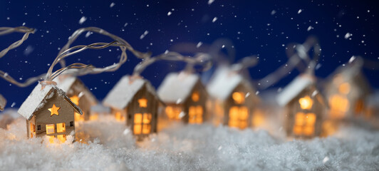 Abstract Christmas Winter Panorama with Wooden Houses Christmas String Lights in Cold Snow Landscape and Glowing Golden Lights in Background. Panorama, Banner. Christmas or Energy themes. - 551763543