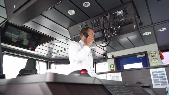 Ships captain talking in vhf radio in modern wheelhouse - Static low angle clip with blurred navigation console in foreground and male captain in middle of wheelhouse