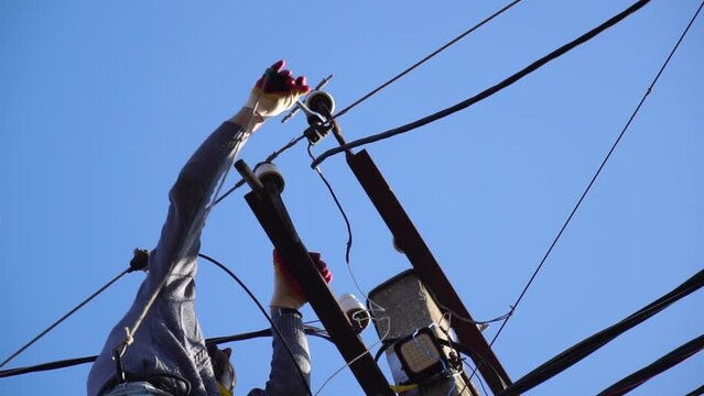 Skilled electrician in helmet fixes wires standing on ladder near high pole against blue sky on summer day backside view. Electrical service and mounting on the pole. Slow motion