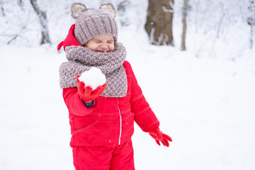 Cute little girl in pink snowsuit and knitted hat and scarf plays with snow in winter forest