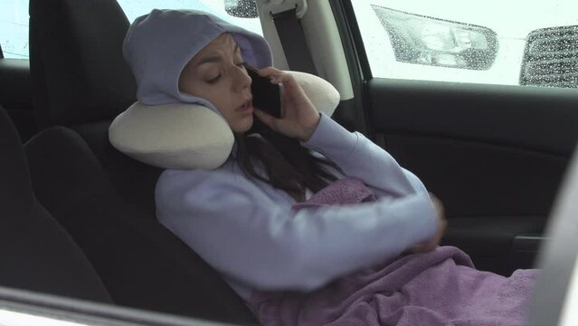 a woman in a neck pillow is resting in the car and talking on phone. wants to sleep. Road trip concept.