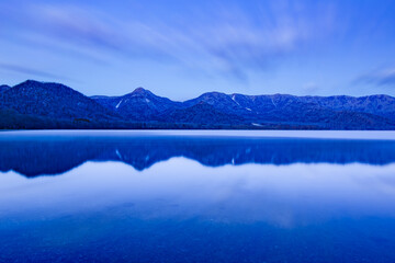 Fototapeta na wymiar 淡いブルーに染まる薄明りの湖の水面に映る山と空。