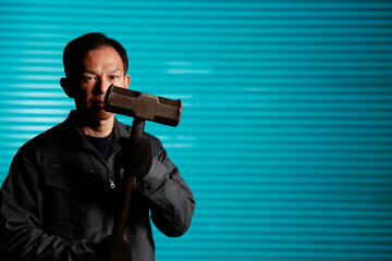 Fototapeta na wymiar Craftsman in flayed work clothes holding a large metal hammer in front of the blue shutters of a town factory. Concept images of the essence of manufacturing and technical succession.