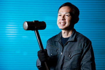 Craftsman in flayed work clothes holding a large metal hammer in front of the blue shutters of a town factory. Concept images of the essence of manufacturing and technical succession.