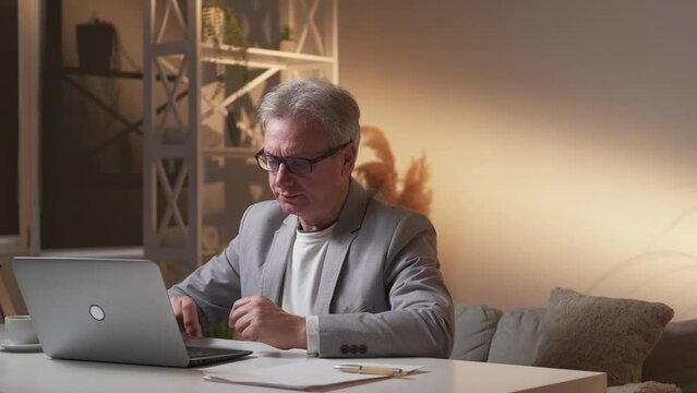 Late work. Smart aged man. Online technology. Worried busy male typing laptop working with documents sitting desk in light shadow room interior.