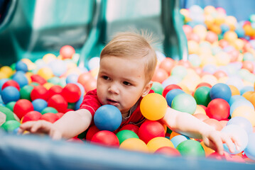 Fototapeta na wymiar Happy laughing boy 1-2 years old having fun in ball pit in kids amusement park and indoor play center. Activity toys for little kid