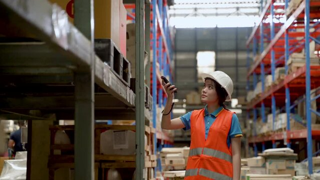 4K, Asian female employee at a large warehouse checks the number of products on the shelves using a barcode scanner to scan the crates of goods. A woman wearing a safety shirt and a safety helmet