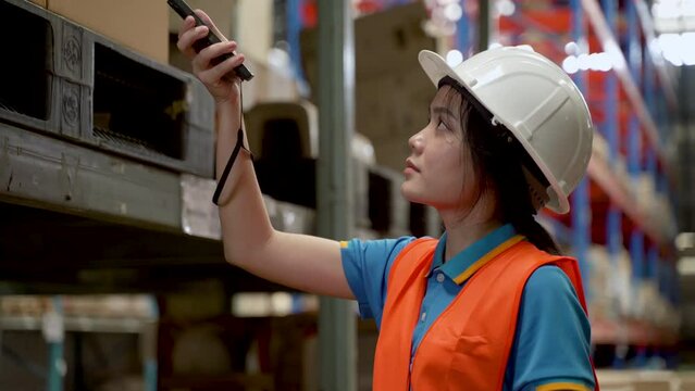 4K, close-up Asian female employee at a large warehouse checks the number of products on the shelves using a barcode scanner to scan the crates of goods. woman wearing safety shirt and safety helmet