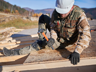 Obraz premium Carpenter hammering nail into OSB panel on the roof top of future cottage. Man worker building wooden frame house. Carpentry and construction concept.