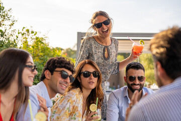 group of fashionable people drinking cocktails, young couples laughing and having fun during an aperitif, travel and luxury holiday concept, focus on blonde smiling woman standing