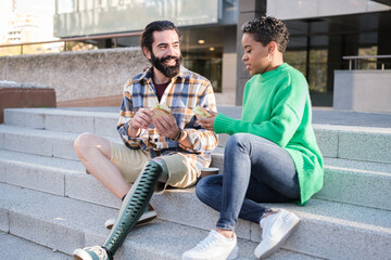 Young man with prosthetic leg and young woman with short hair having a fun time eating a sandwich outdoors