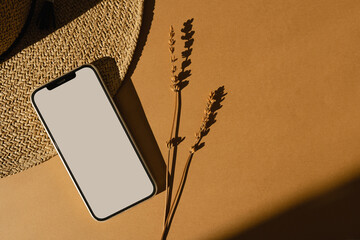 Smartphone with blank mockup copyspace screen, straw hat, dried grass stems on warm tan background...