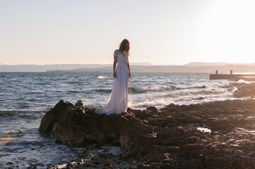 Beautiful young bride in a white wedding dress stands on the ocean or sea in the sun. Fashion shooting in a wedding or evening dress